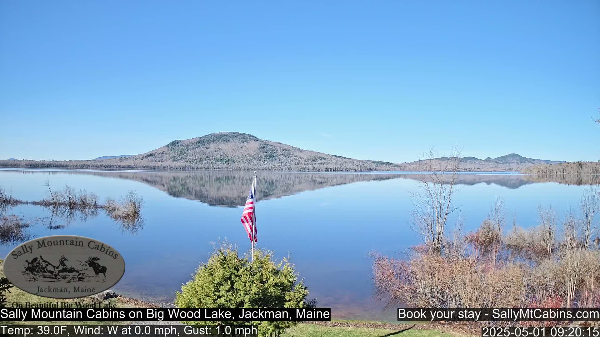 A calm, clear day reveals a tranquil lake reflecting a distant mountain range, with an American flag standing in the foreground.