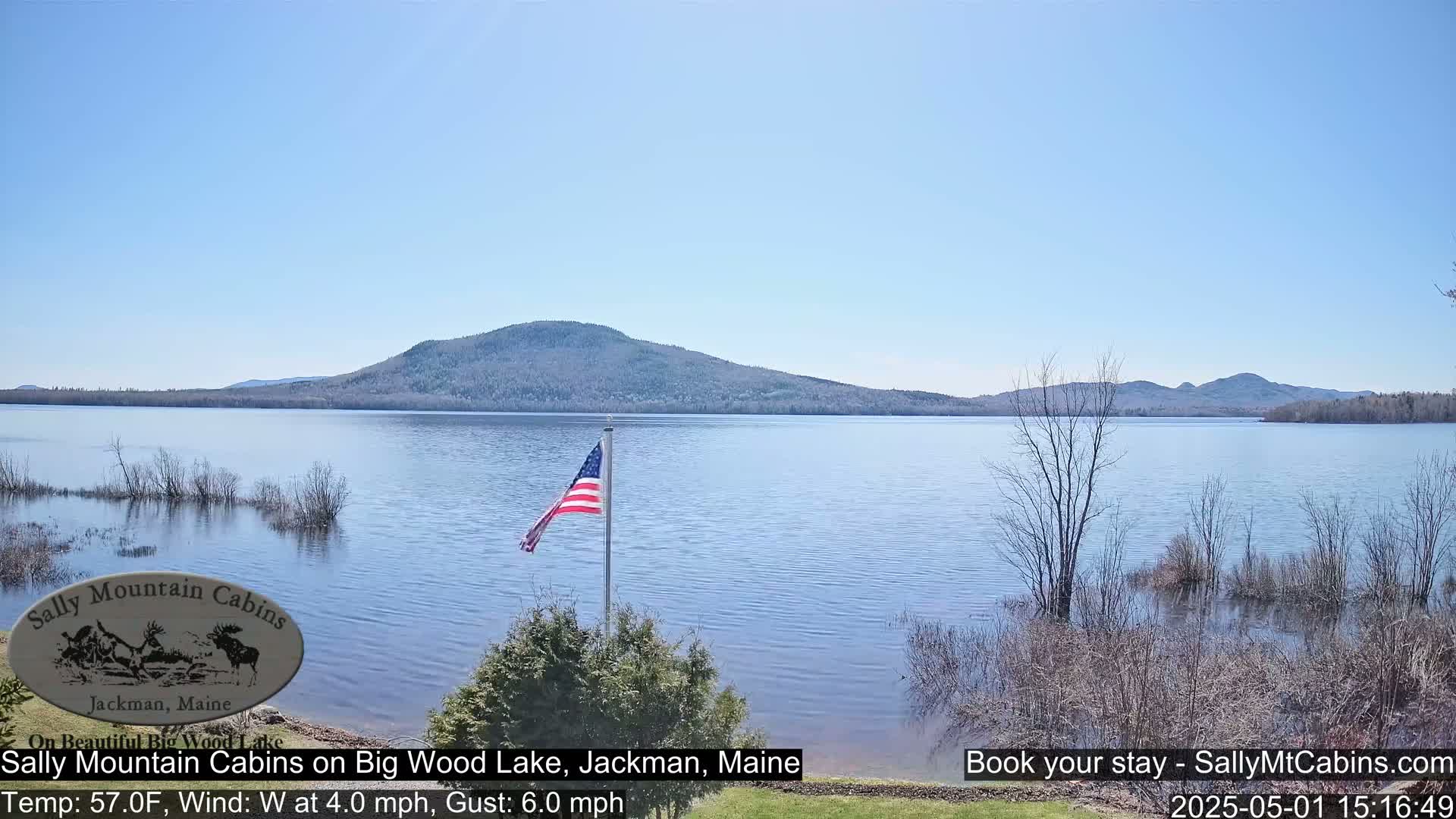 A calm lake under a clear blue sky is backed by a mountain range and a US flag on a pole in the foreground.
