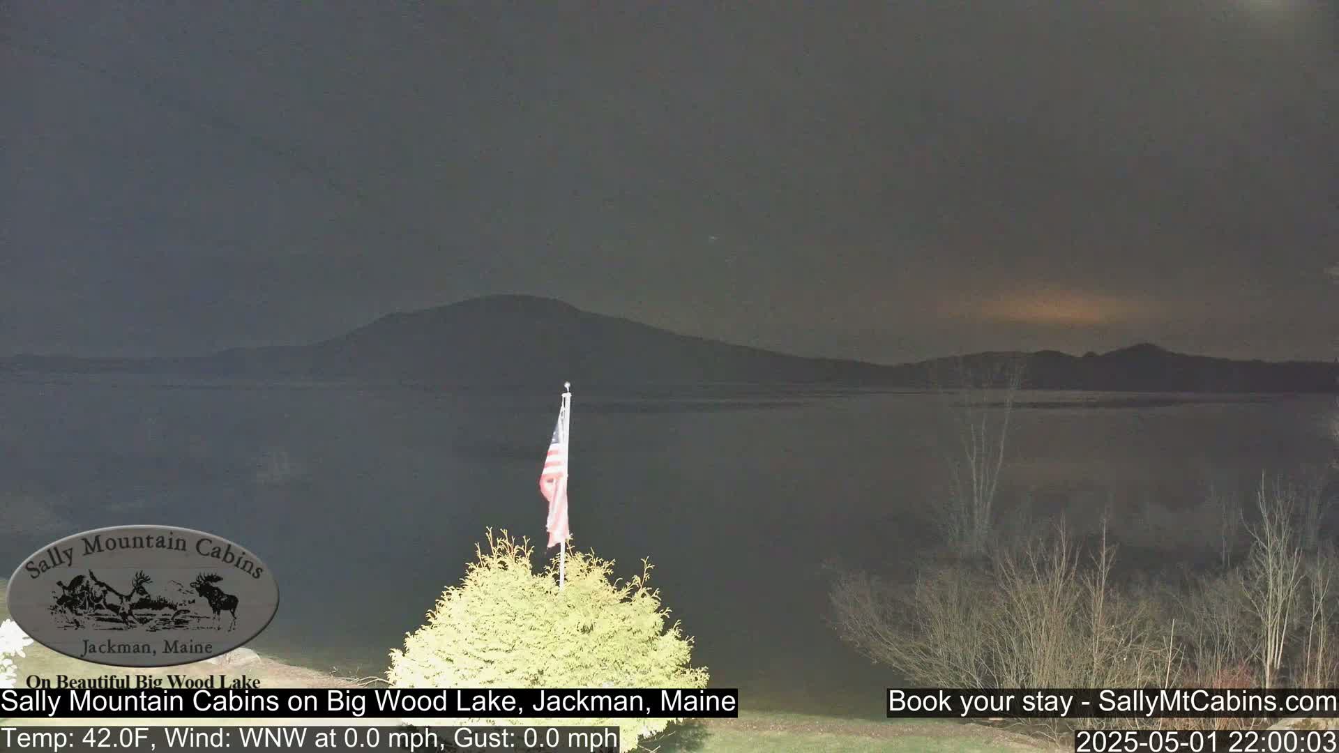 A calm, dark night scene shows a lake with a mountain in the background and an American flag on a pole in the foreground.