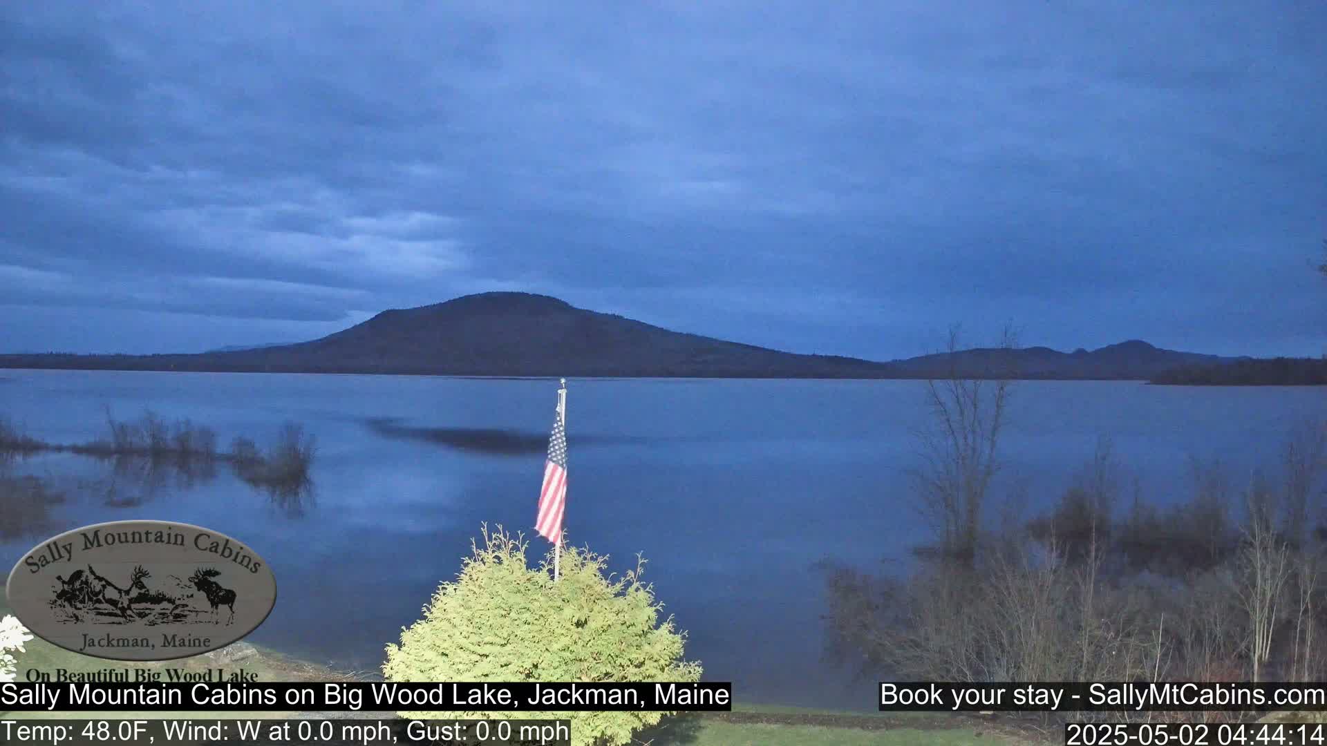 A calm, dark-blue lake is viewed from a lakeshore, under a twilight sky, with a small American flag planted in the foreground.
