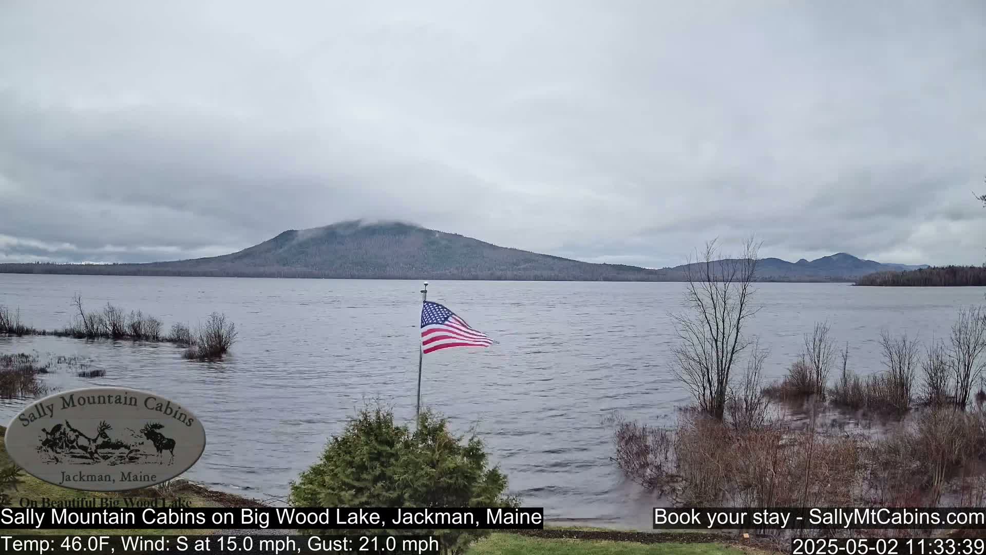 An American flag waves on a lakeshore under a cloudy sky, with a mountain range visible in the distance and the water appearing somewhat flooded.