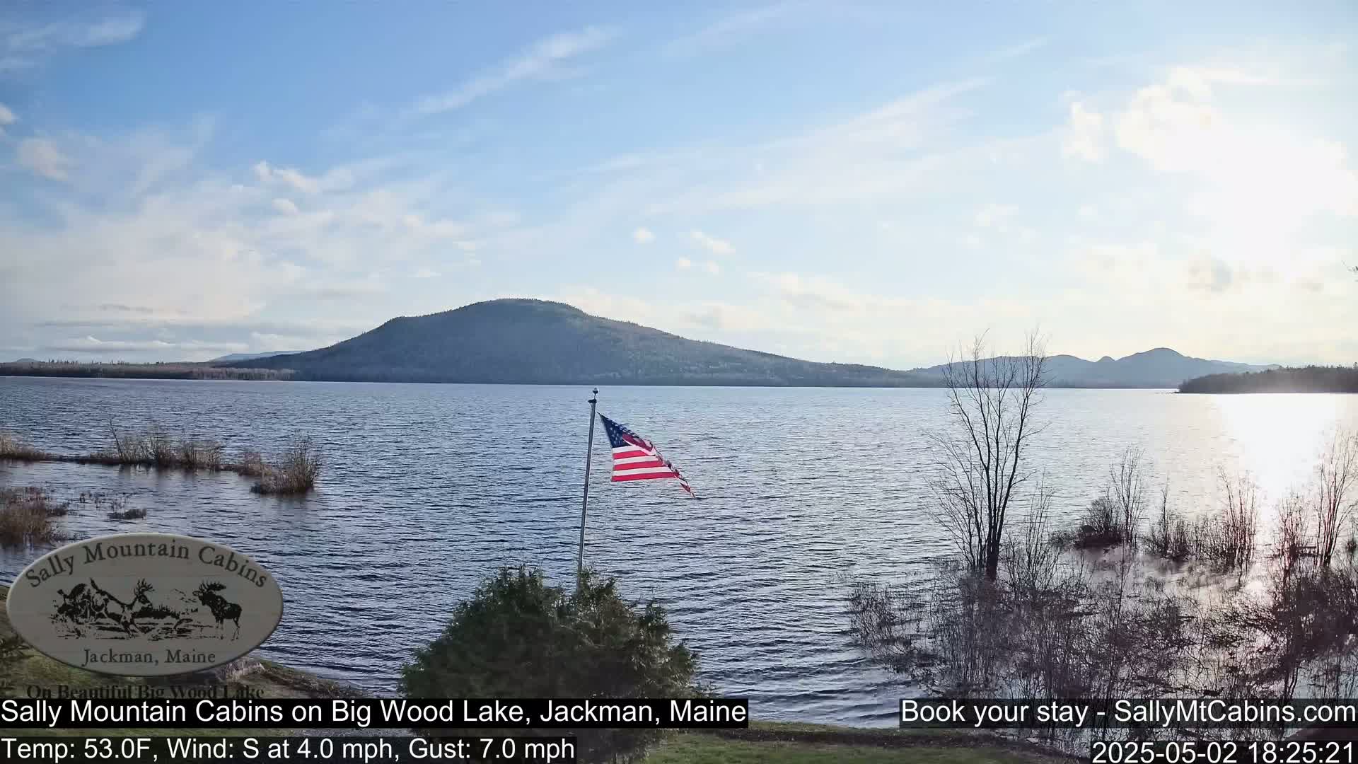 An American flag flies near a calm lake under a partly cloudy sky, with mountains in the distance.
