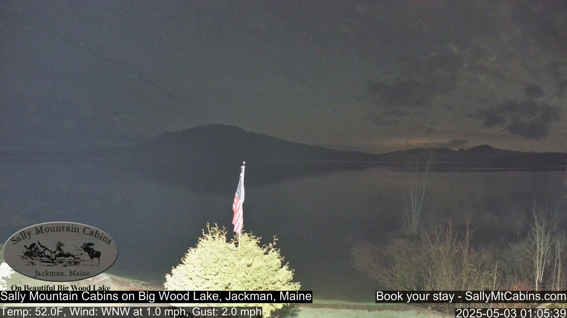 A nighttime view of a calm lake under a partly cloudy sky, with a small American flag visible in the foreground.