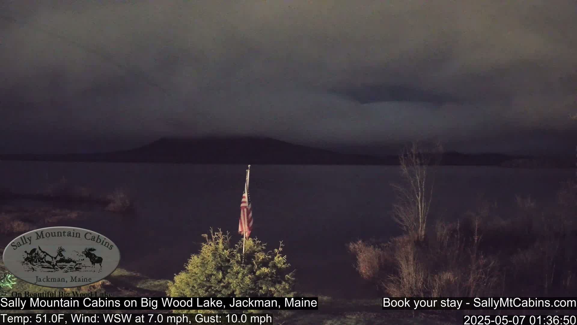 A dark, overcast night scene shows a calm lake with a distant mountain range under a mostly cloudy sky; a small American flag stands in the foreground near some low shrubs.