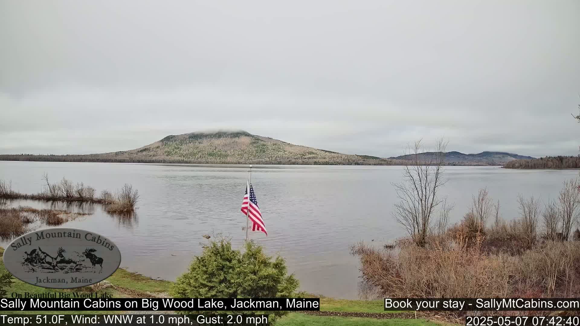 A calm lake with a hazy mountain in the background under an overcast sky, an American flag stands in the foreground.