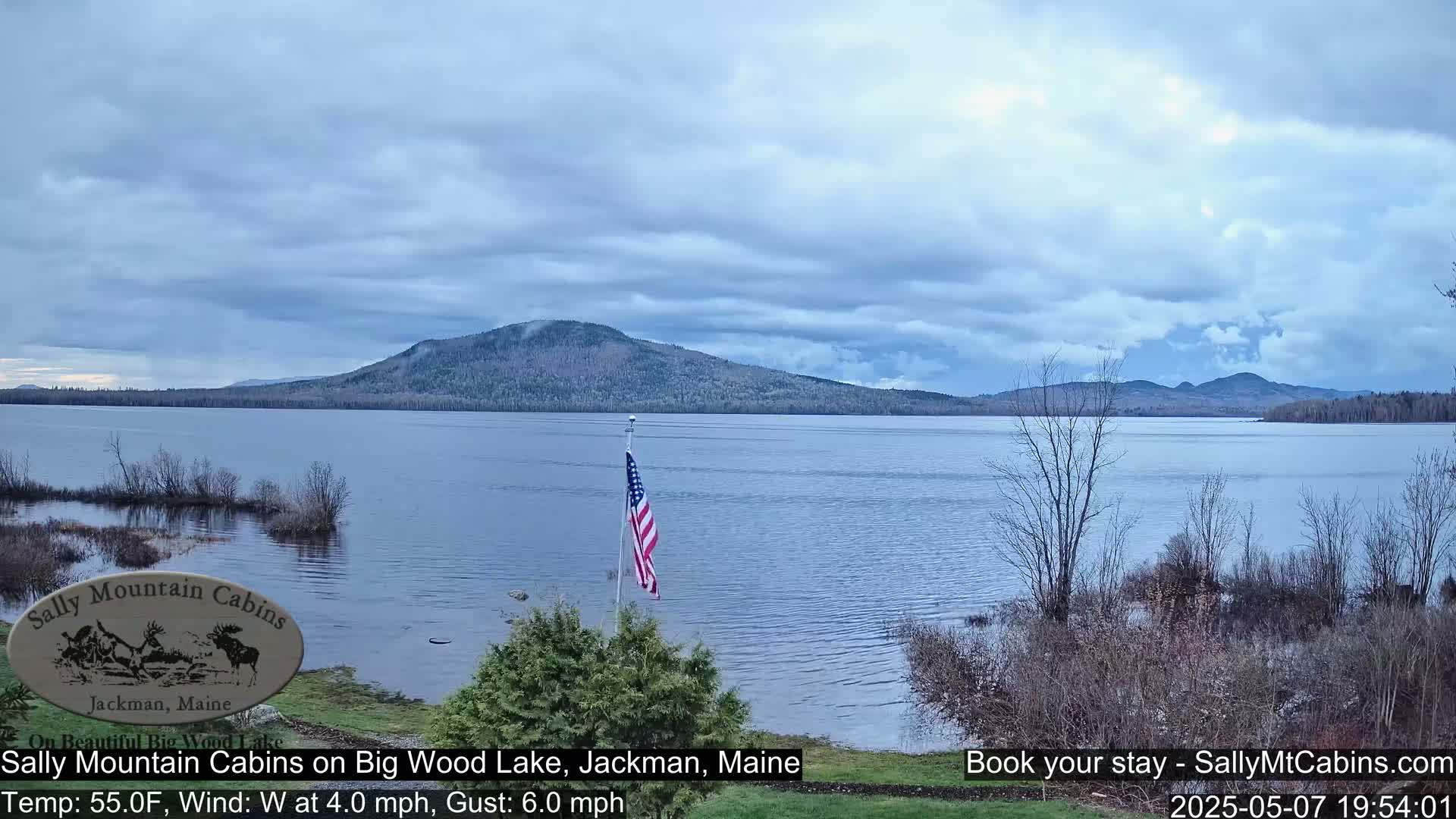 An American flag stands near a lake under a mostly cloudy sky, with a mountain range in the distance.
