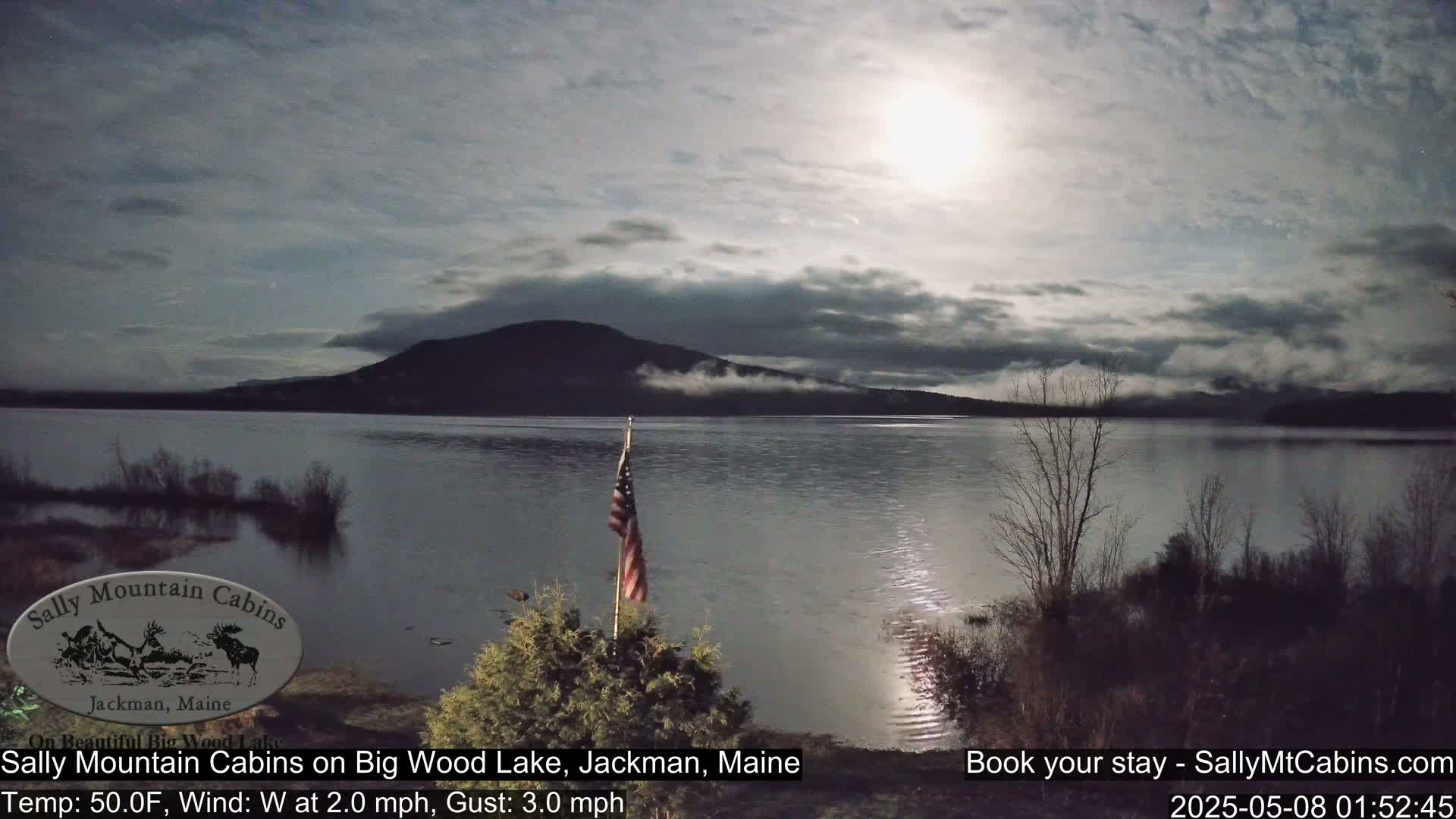 A calm lake under a mostly cloudy night sky, illuminated by a bright moon, with a small American flag standing near the water's edge.