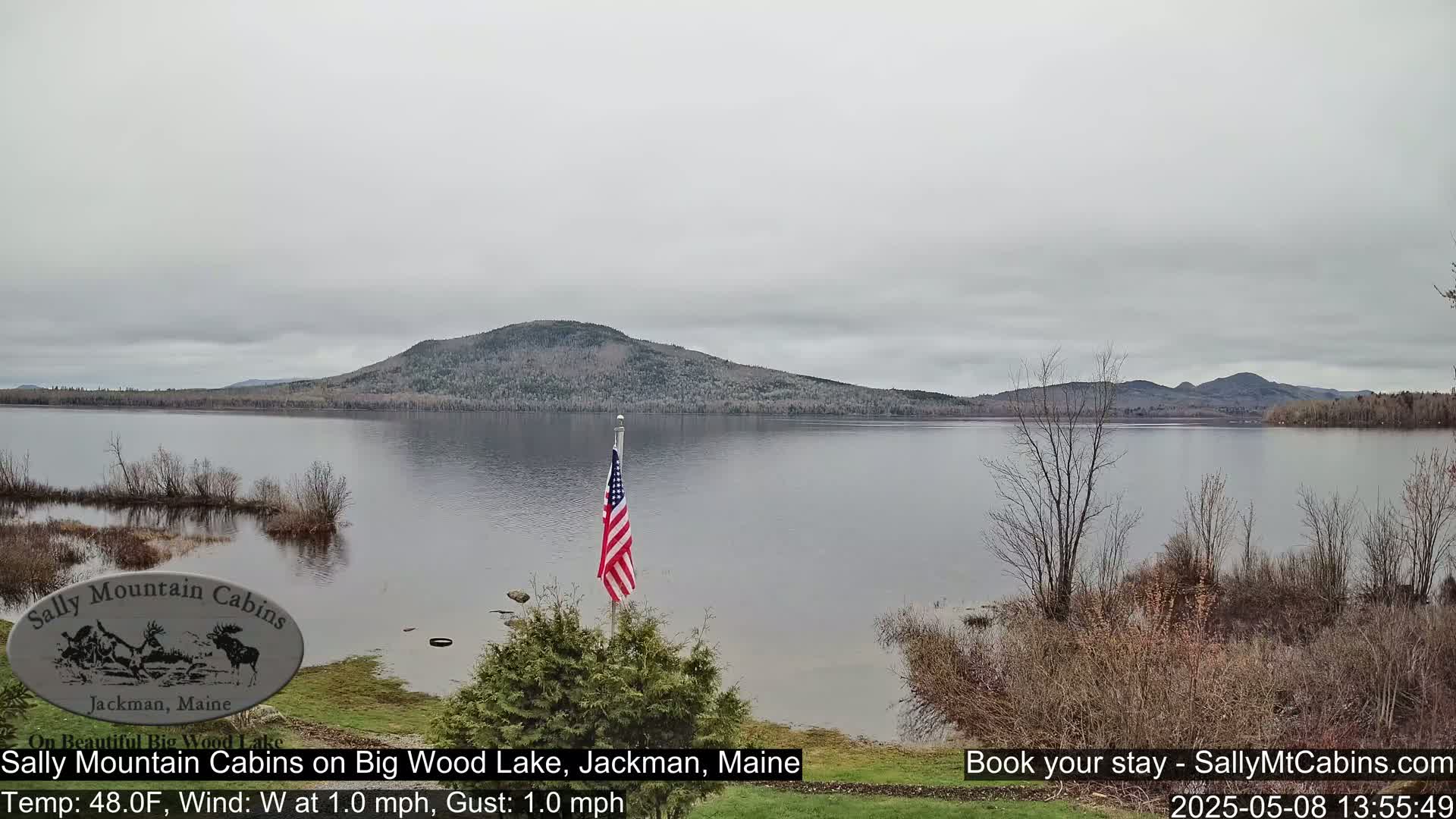 A calm, overcast day shows a view of a lake with a forested mountain in the background and an American flag in the foreground.