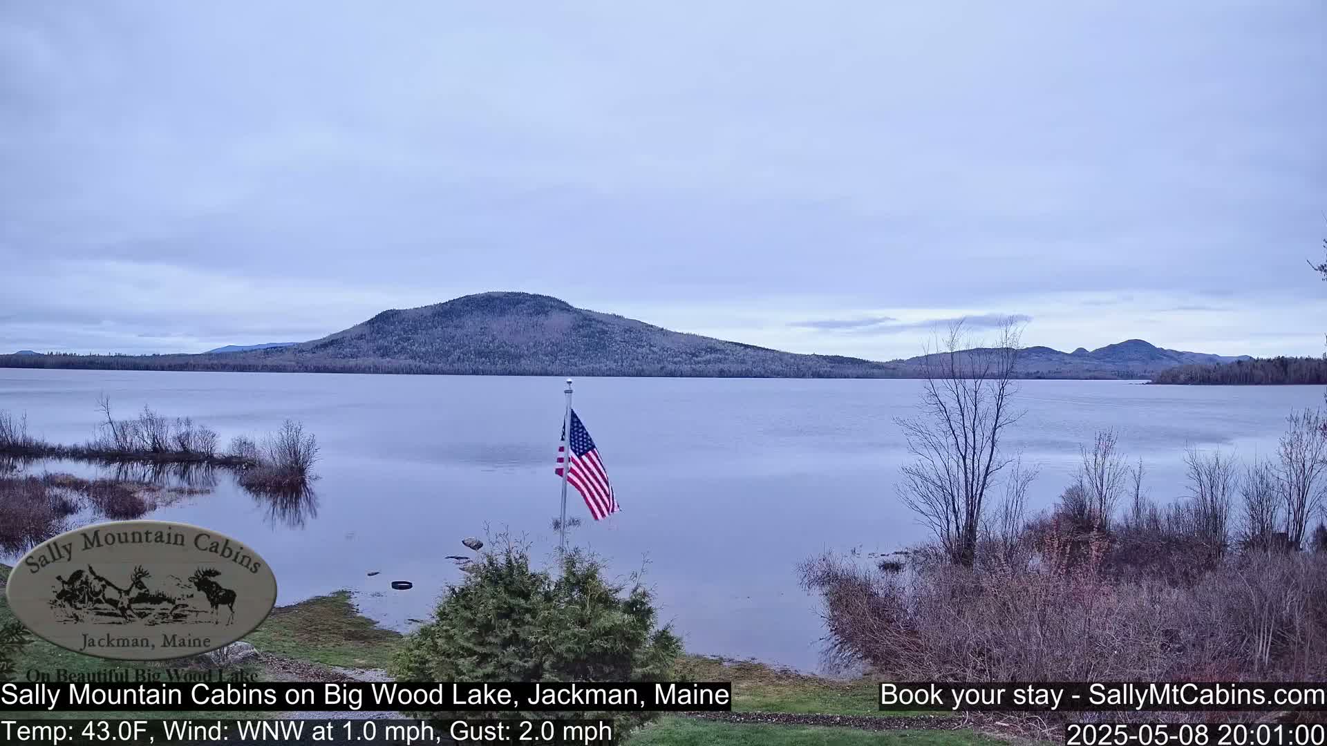 A calm lake with a distant mountain range is visible under a cloudy sky, and an American flag is flying near the shoreline.