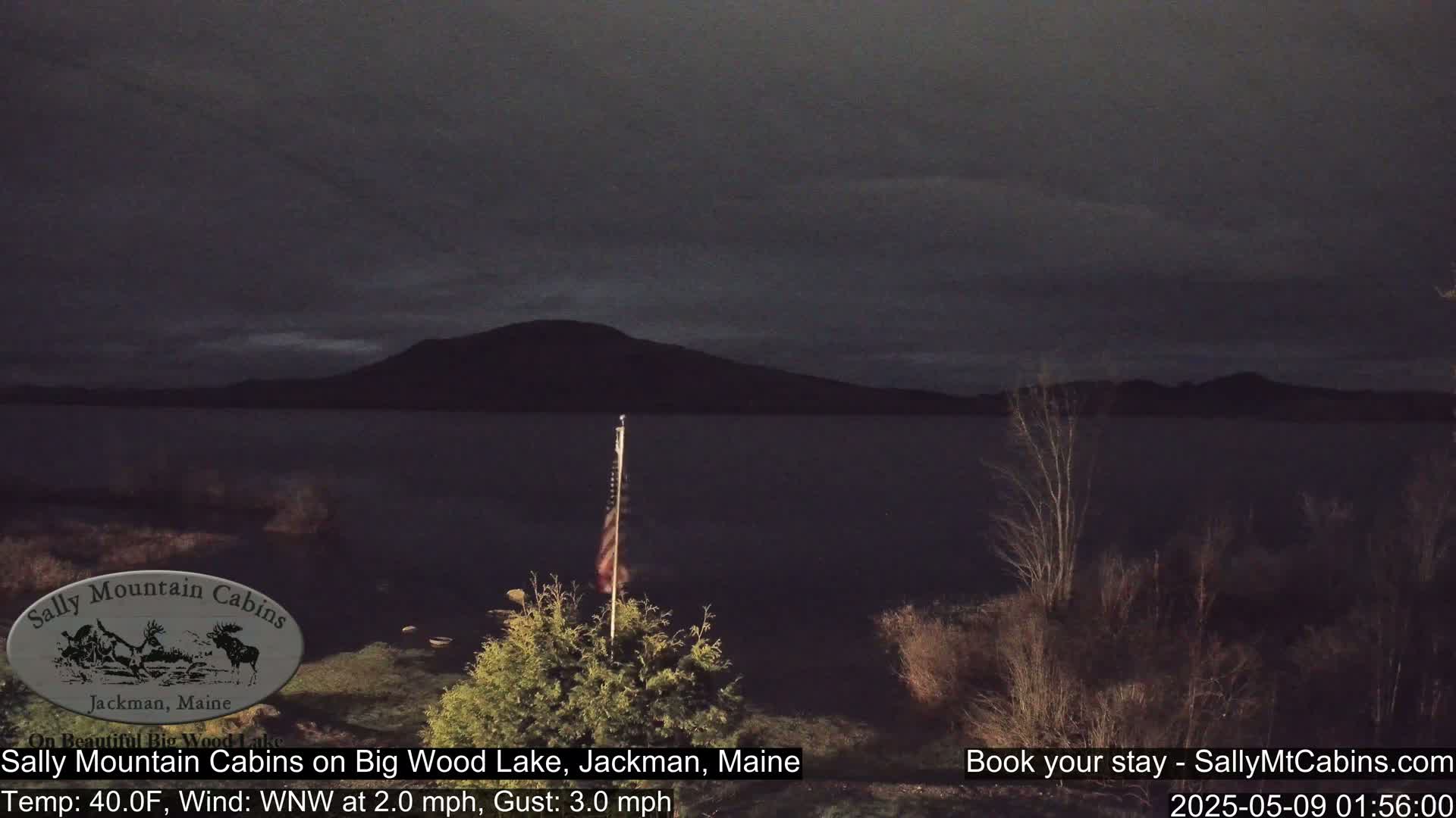 A dark, calm lake is seen at night under a mostly cloudy sky, with a small American flag visible in the foreground.
