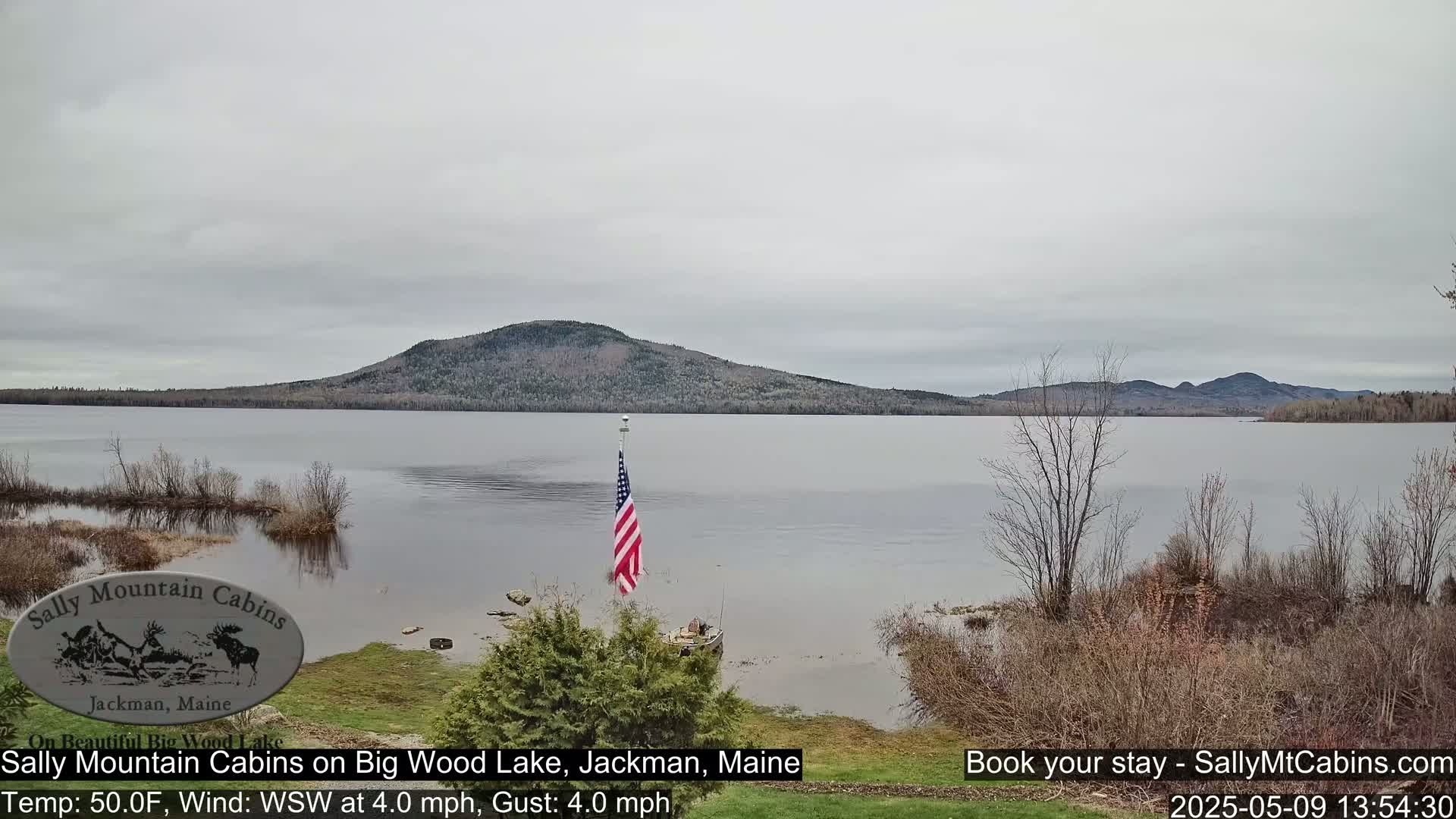 A calm lake under an overcast sky is seen with a distant mountain range and an American flag on a flagpole near a small boat on the lakeshore.