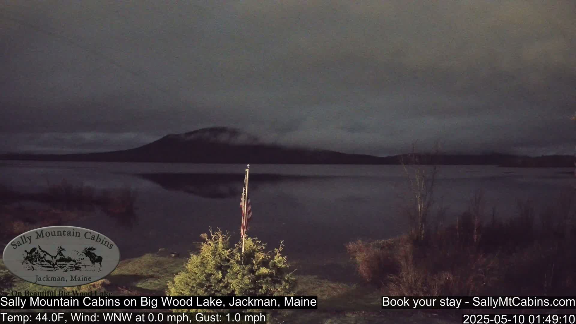 A calm, dark lake is seen under a gray, overcast sky, with a mountain in the background and an American flag in the foreground.
