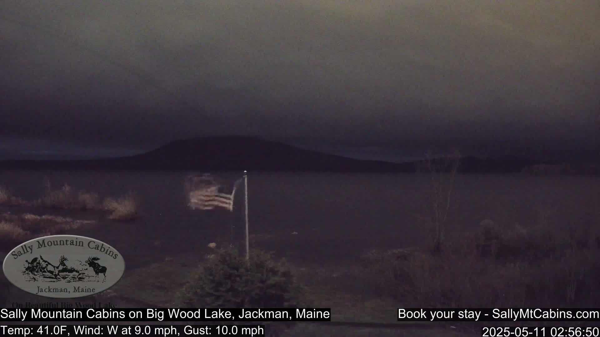 A dark, overcast scene shows a calm lake with a distant shoreline, a flagpole with an American flag waving in a west wind, and sparse vegetation in the foreground.