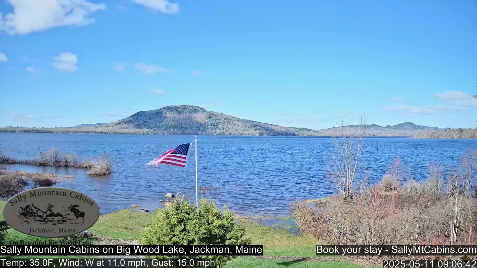 A calm, blue lake with a gently waving American flag is seen from a lakeside vantage point, with a mountain range in the background under a mostly clear sky.