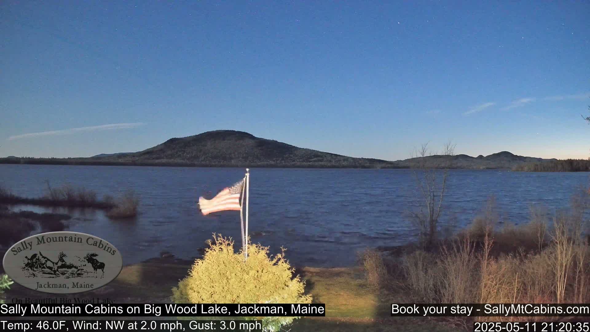 An American flag waves gently near a calm lake under a twilight sky, with a dark mountain in the distance.
