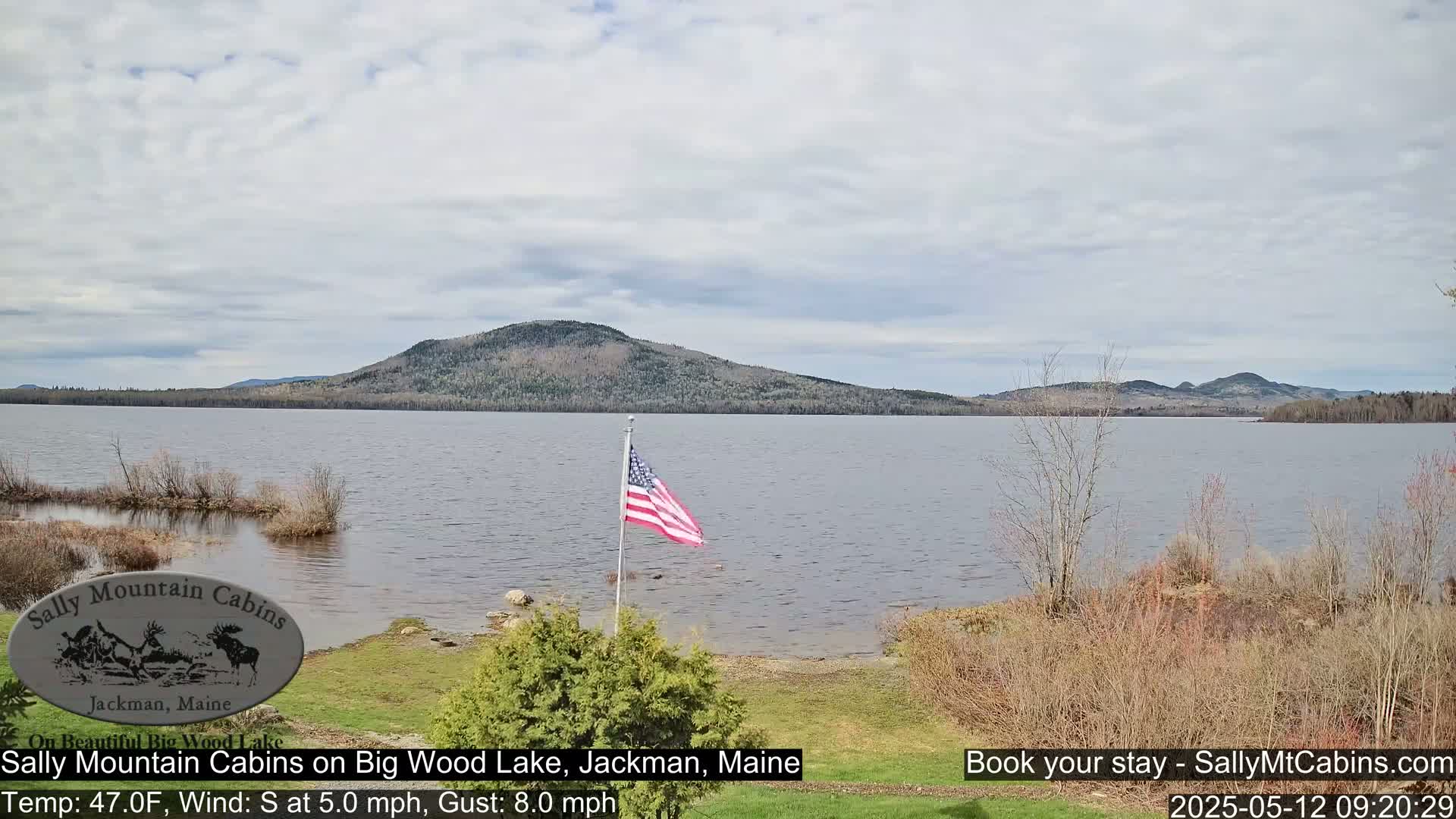 A calm lake with a gently sloping shoreline, a distant mountain range, and an American flag waving in a south wind under a mostly cloudy sky.