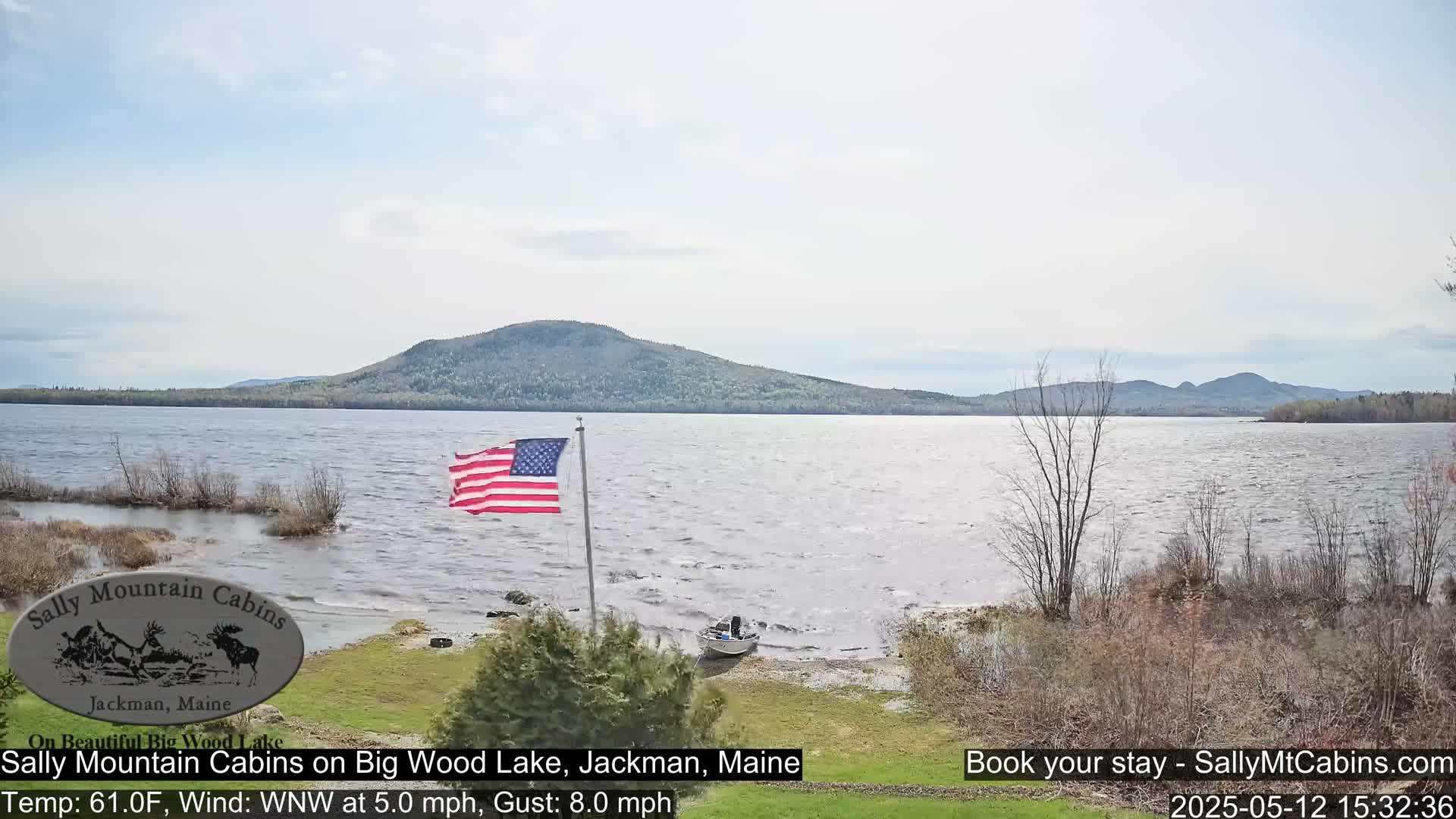 A slightly windy day shows an American flag waving near a small boat on the shore of a lake, with mountains in the background.