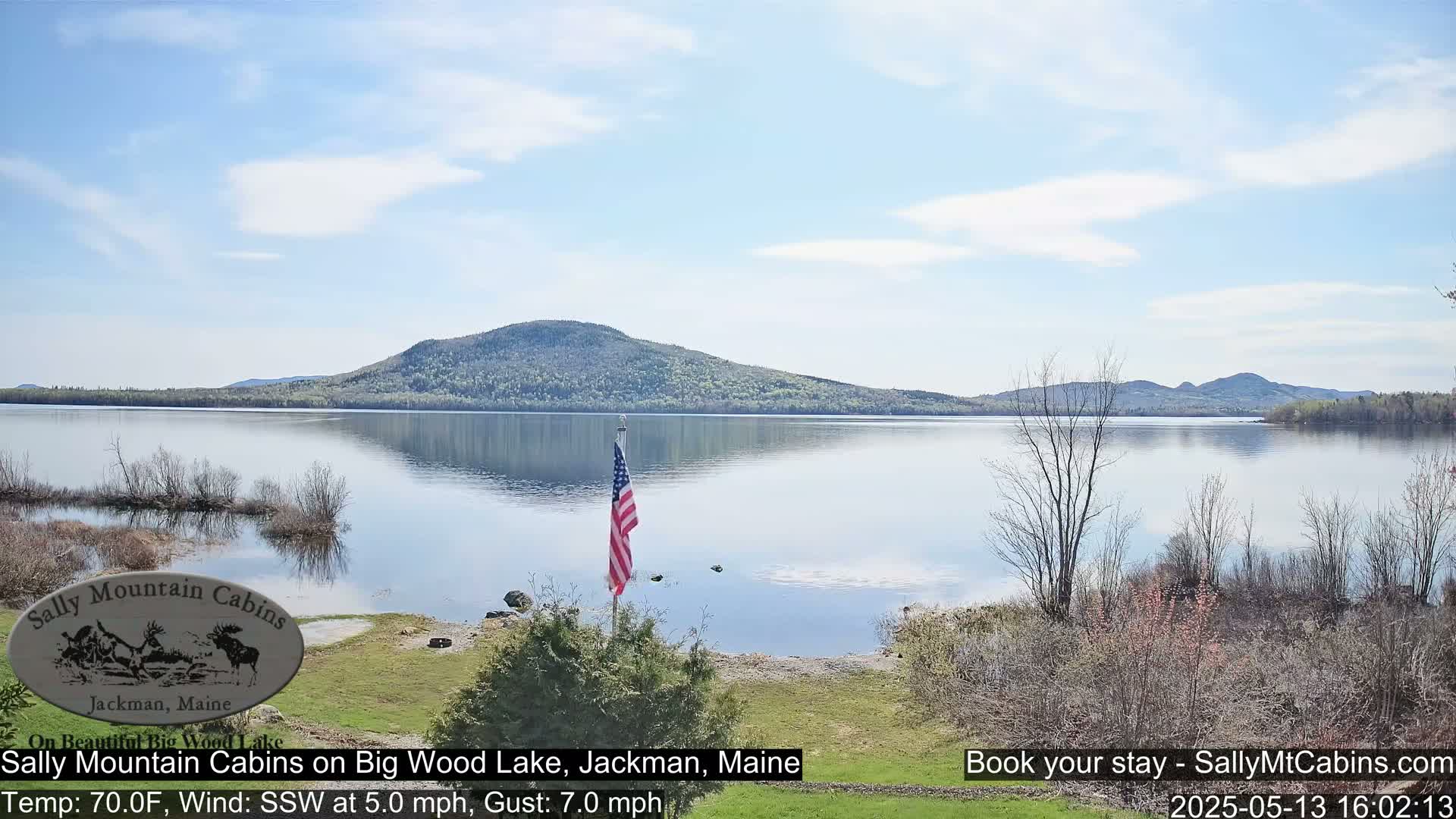 A calm lake with a mountain in the background under a partly cloudy sky, a small American flag is planted near the shoreline.