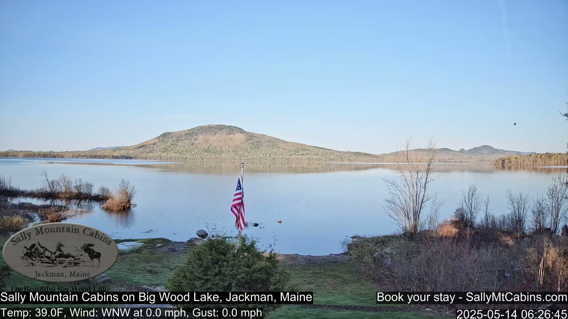 A calm, clear day shows a tranquil lake reflecting a forested hill under a pale blue sky; an American flag stands near the lakeshore.