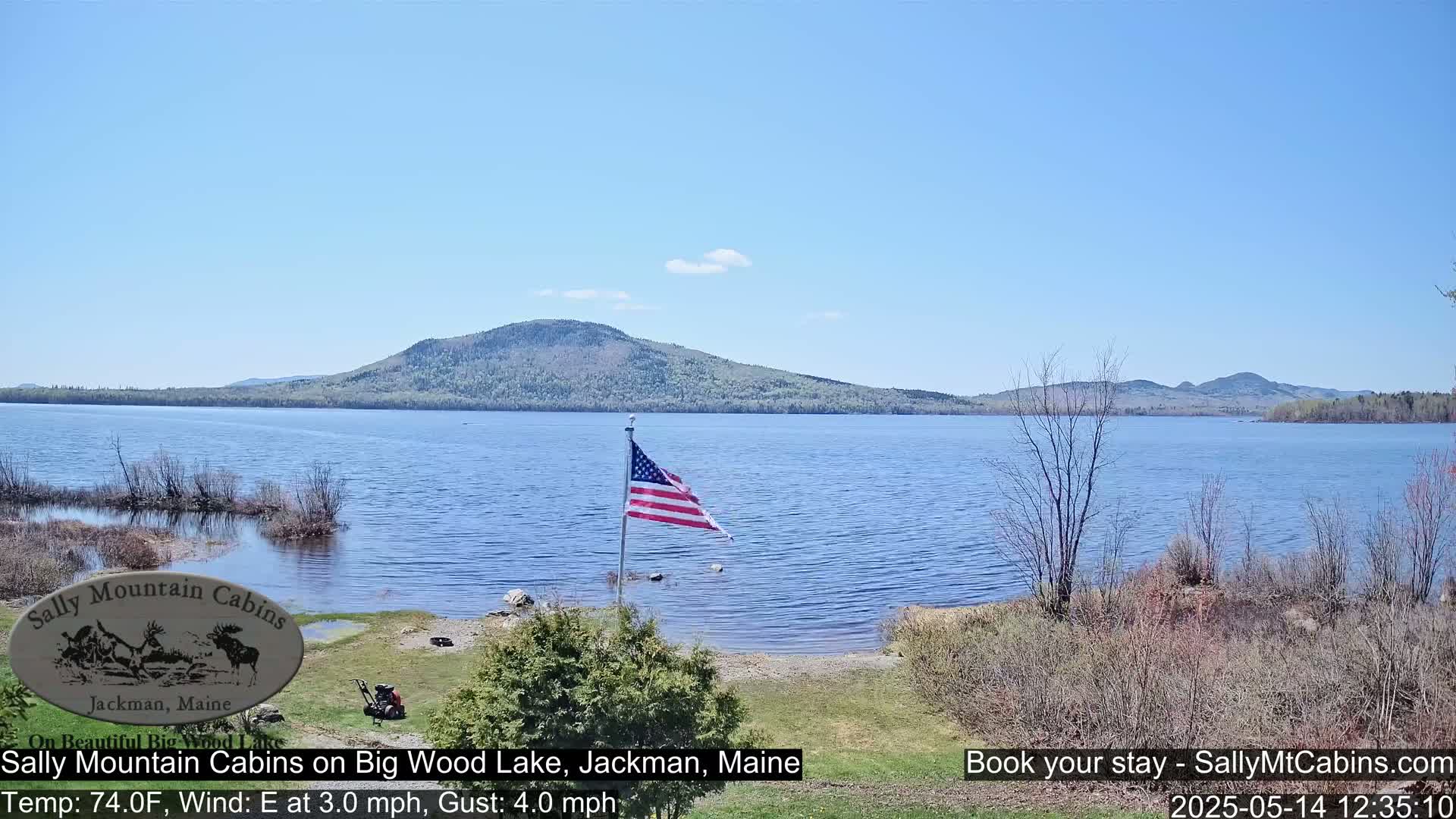 An American flag flies near a calm lake under a clear blue sky, with a forested mountain in the background.