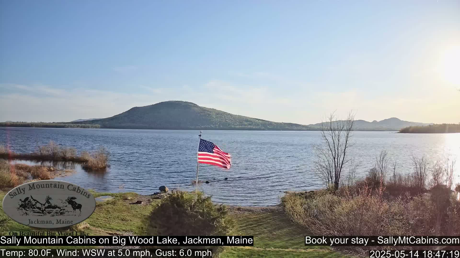 An American flag waves gently near a calm lake under a mostly clear sky, with a mountain range in the background.