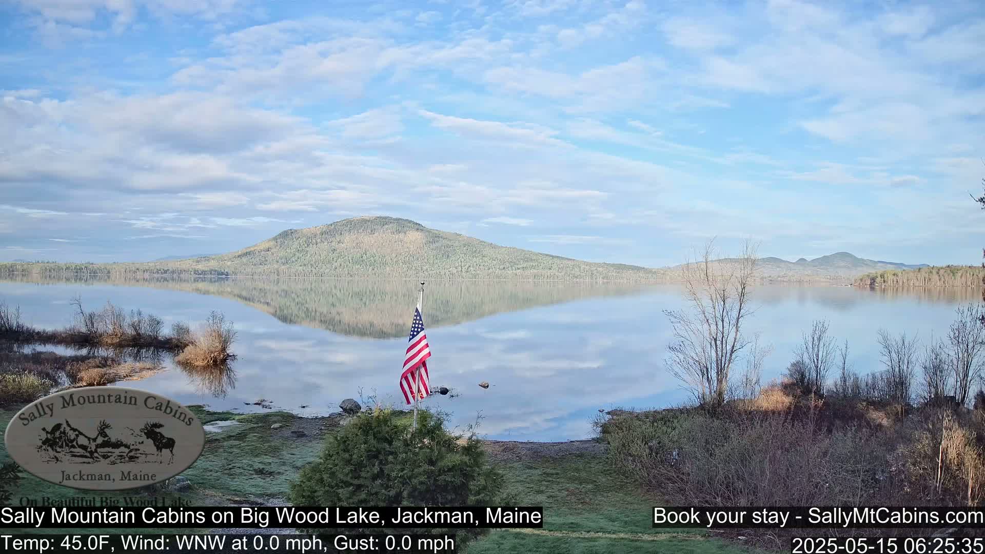A calm lake reflects a treeline and distant mountains under a partly cloudy sky, with an American flag standing near the shore.