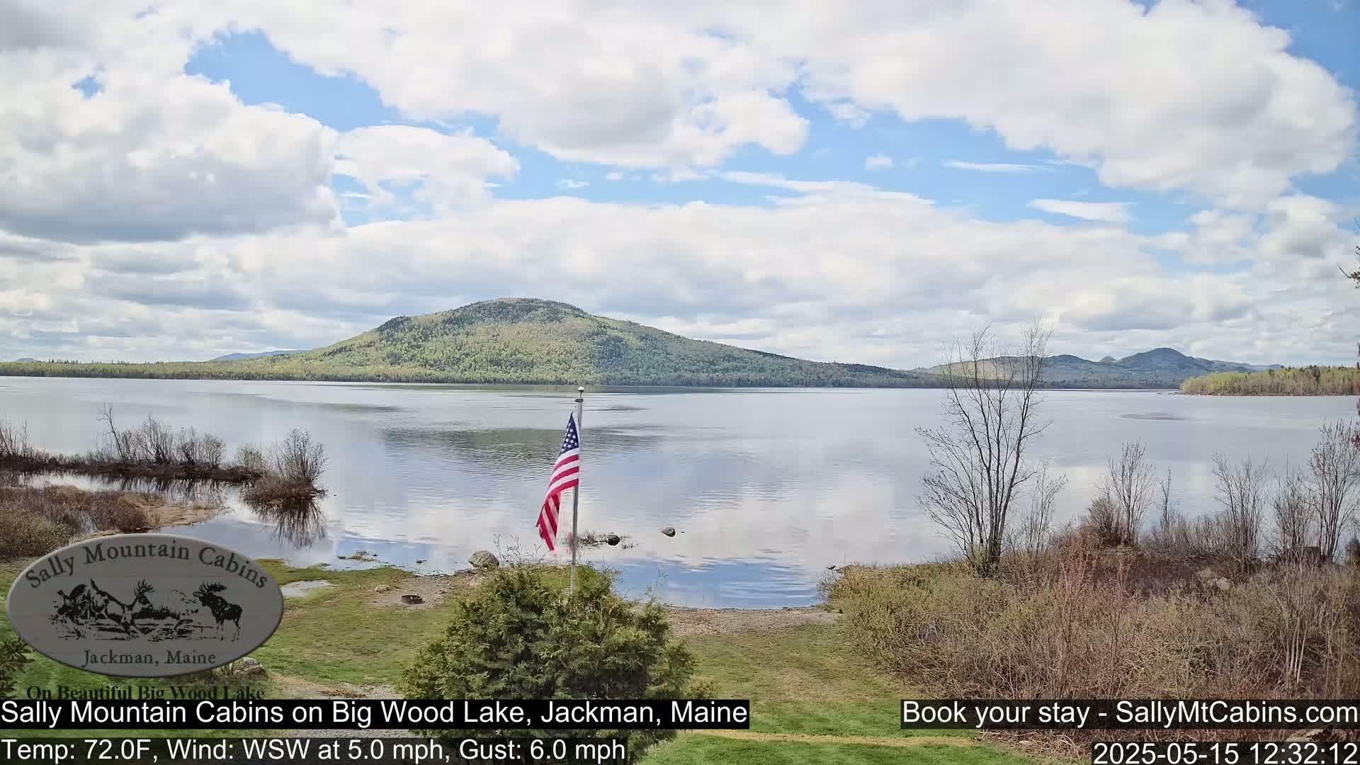 A calm lake with a wooded mountain in the background under a partly cloudy sky, an American flag stands near the shore.