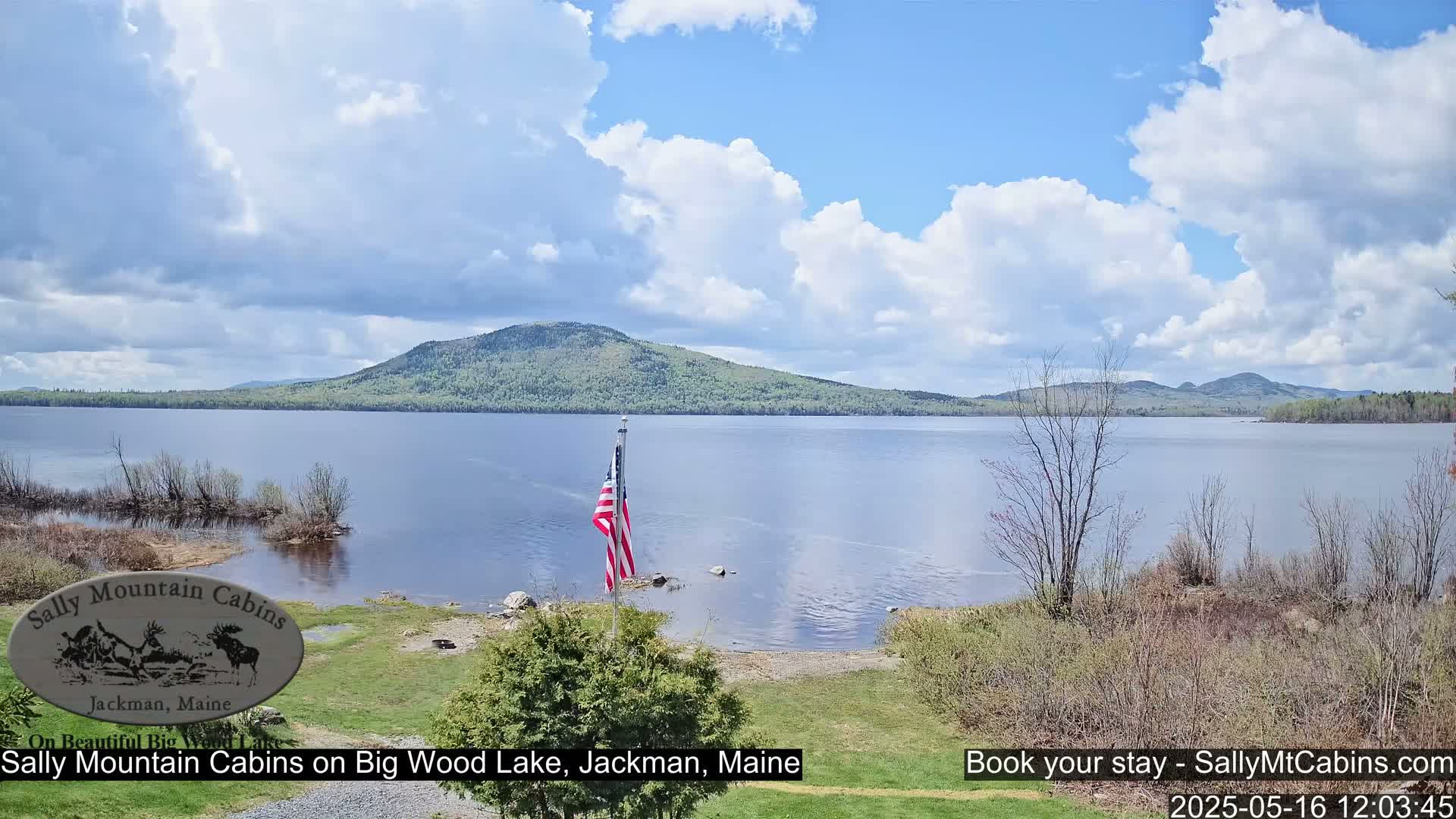 A calm lake with a mountain range in the background under a partly cloudy sky, an American flag stands on the lakeshore.