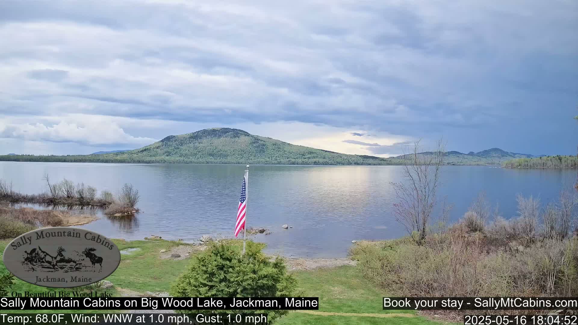 A calm lake with a wooded mountain in the background under a partly cloudy sky is viewed from a grassy shoreline with an American flag.