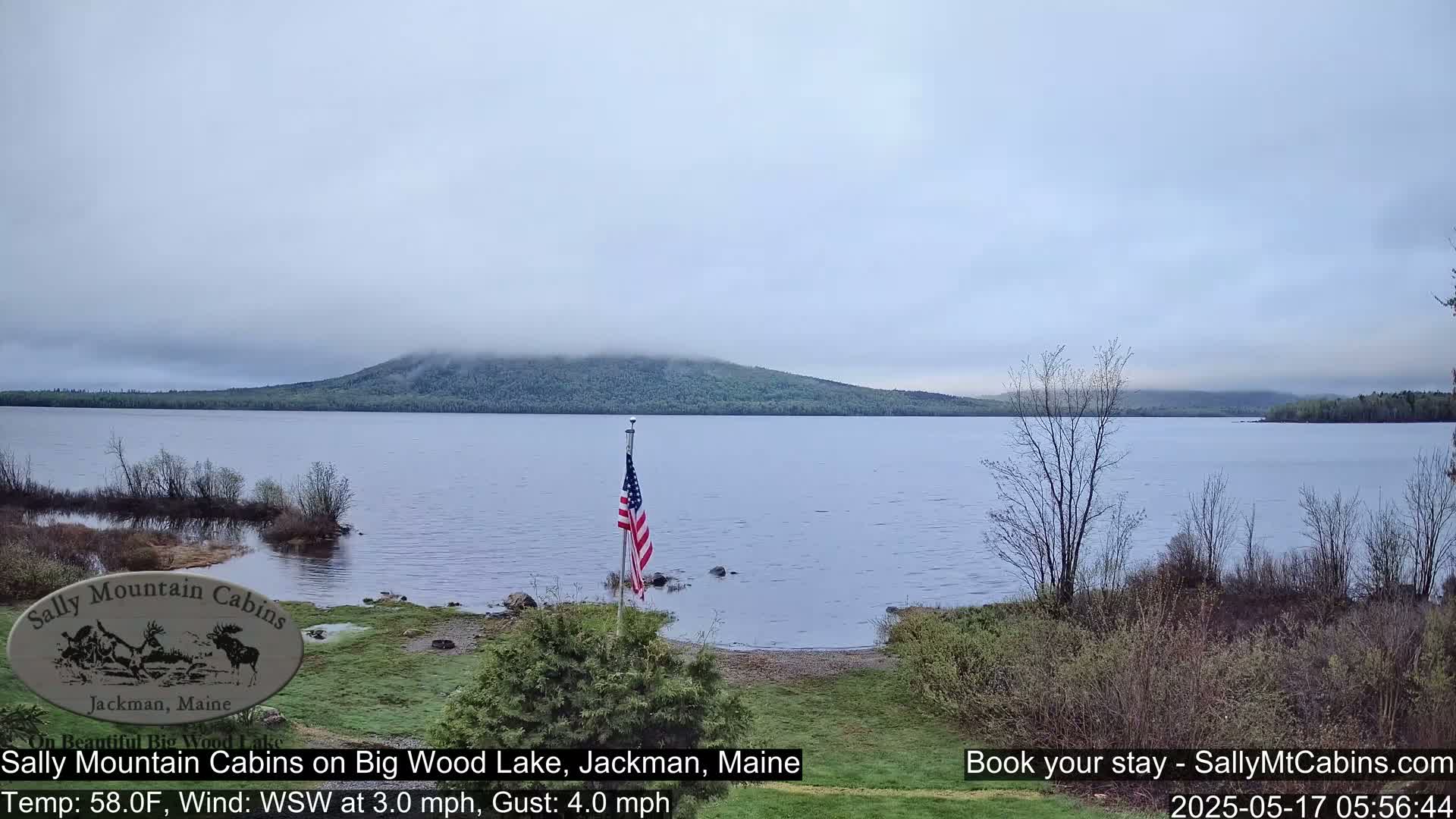 A calm lake with a misty mountain in the background is seen from a lakeside vantage point, with an American flag in the foreground under an overcast sky.