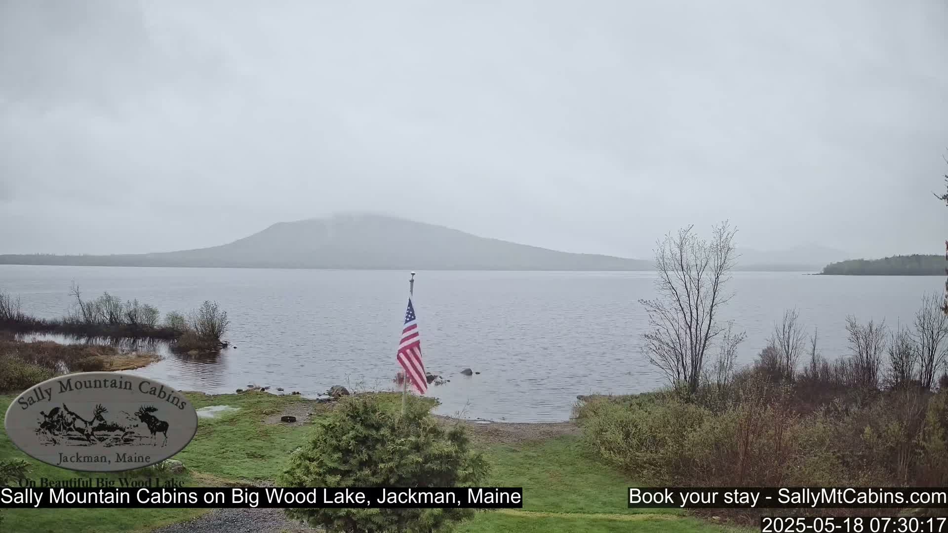 A lake with a hazy mountain in the background is viewed from a lakeside property on a cloudy, possibly rainy day, with an American flag planted on the shore.