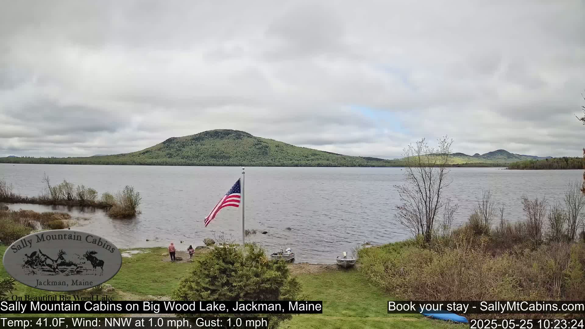 A calm lake under an overcast sky is seen from a lakeshore with two people standing near the water, a flagpole flying an American flag, and several small boats pulled ashore.