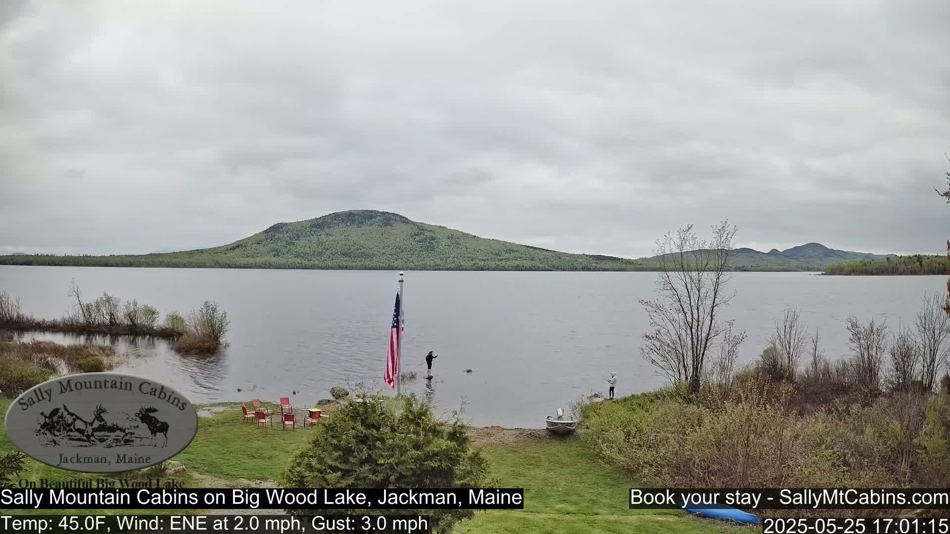 A calm lake under an overcast sky is bordered by green vegetation and a forested hill, with people fishing near a small boat and American flag.