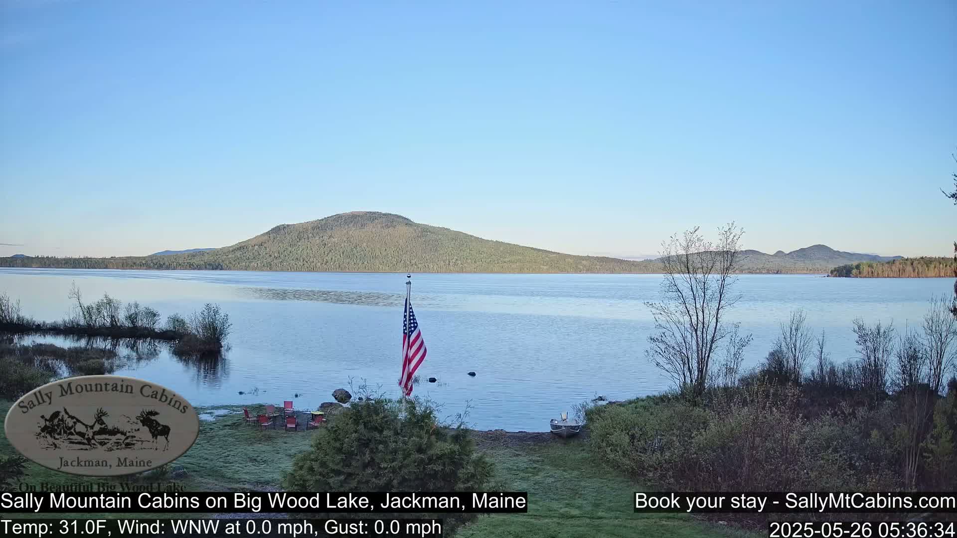 A calm lake with a forested mountain in the background is viewed from a lakeside location with a few chairs and an American flag; the sky is clear and blue.