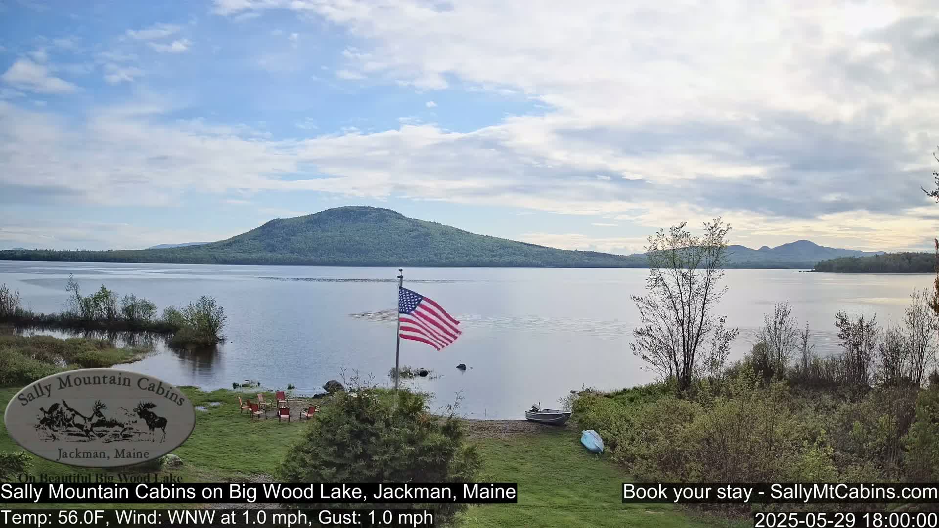 A calm lake with a gently sloping wooded hill in the background under a partly cloudy sky, viewed from a lakeside area with a small American flag.