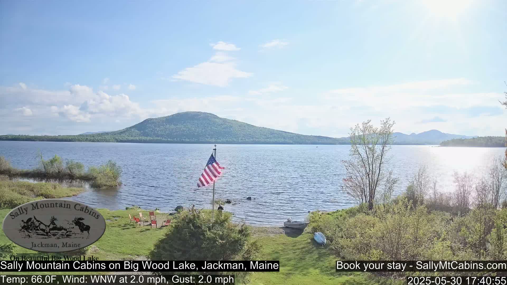 A calm lake with a gently sloping shoreline, a small American flag, and a few chairs sits in front of a forested mountain range under a partly cloudy sky.