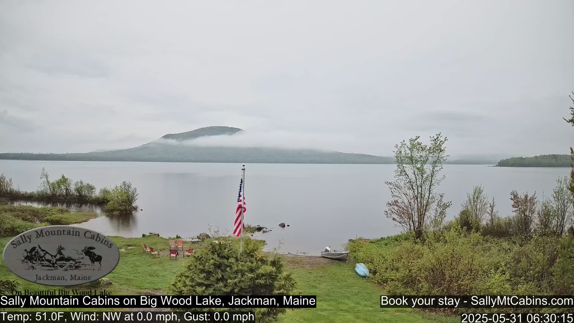 A calm lake with a misty mountain in the background is viewed from a lakeside area with a flagpole, several chairs arranged in a circle, and a small boat.