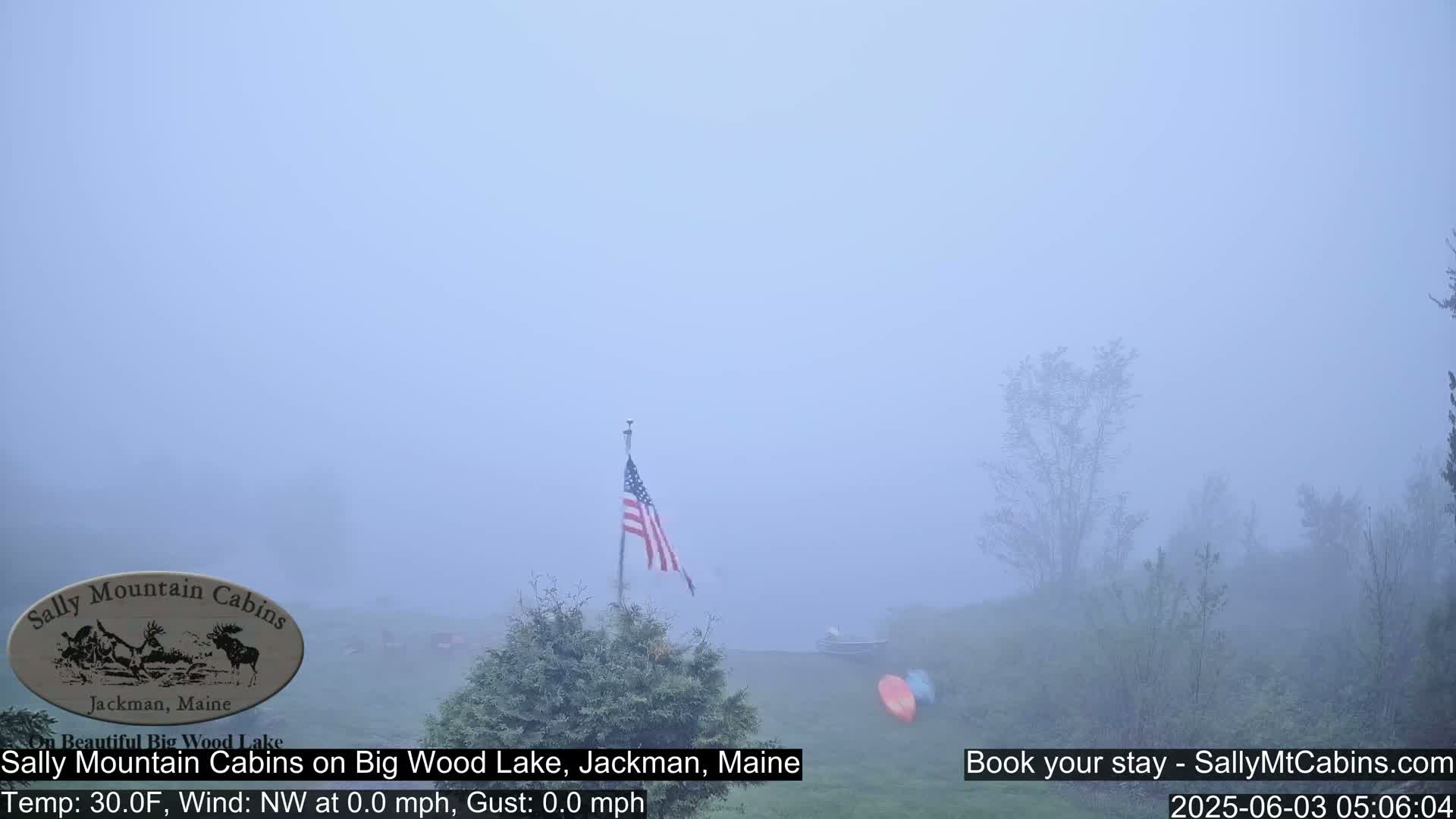 A foggy lakeside scene shows an American flag on a pole, some trees, and a glimpse of a body of water with two kayaks.