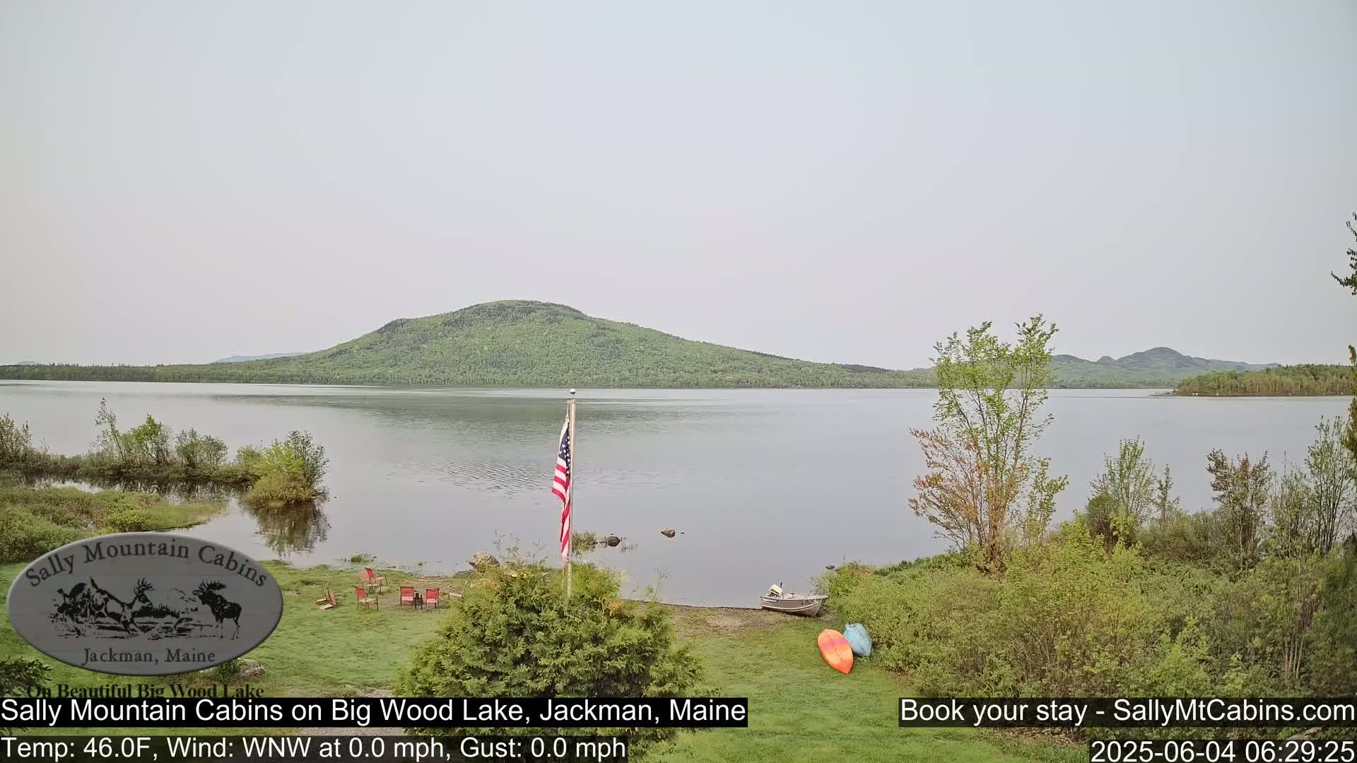 A calm lake with a green, tree-covered hill in the background, under an overcast sky, is viewed from a lakeside lawn with a few chairs and kayaks visible.
