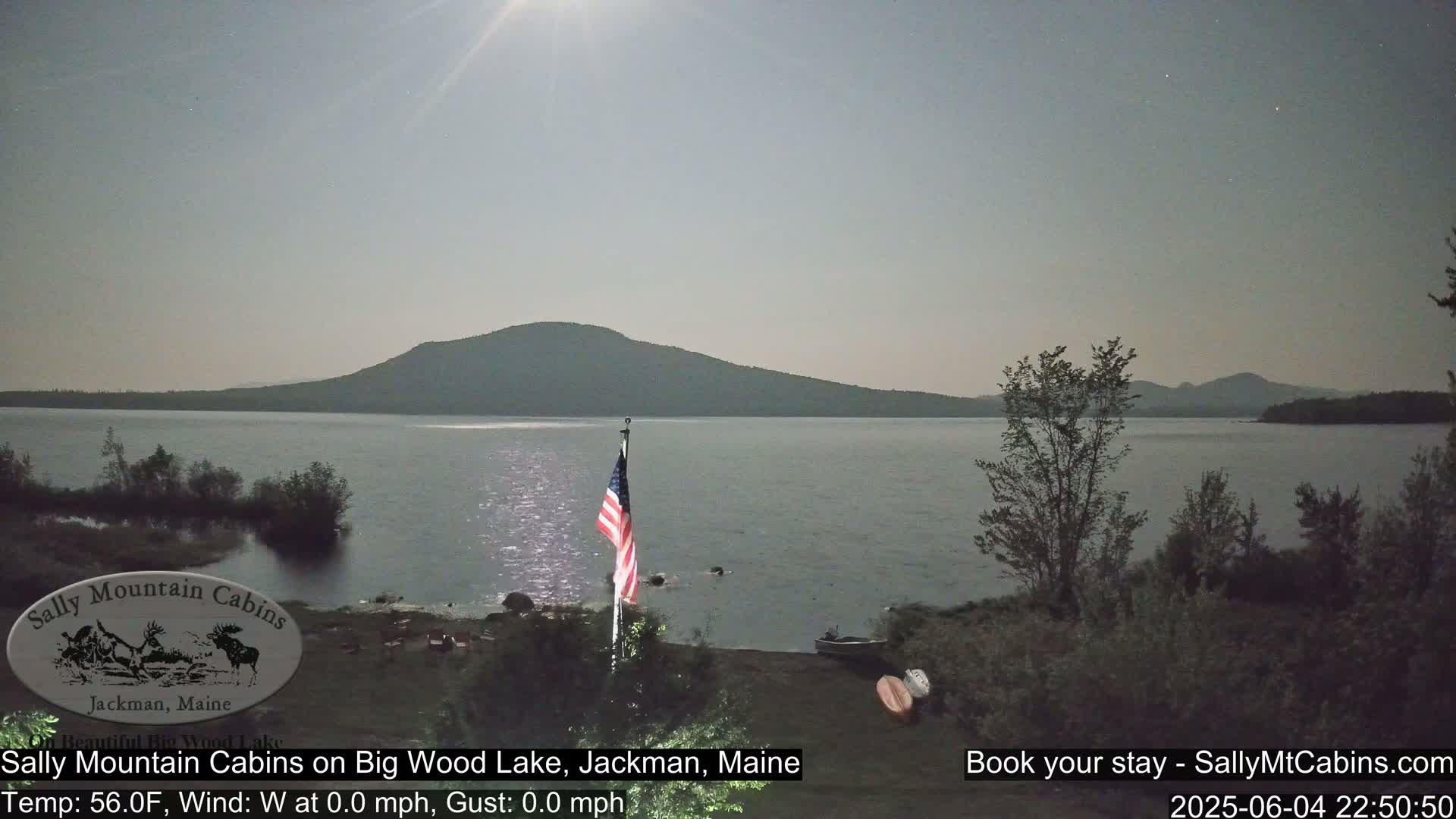 A calm, nighttime scene shows a lake under a bright moon, with a mountain in the background and an American flag standing near the shore.