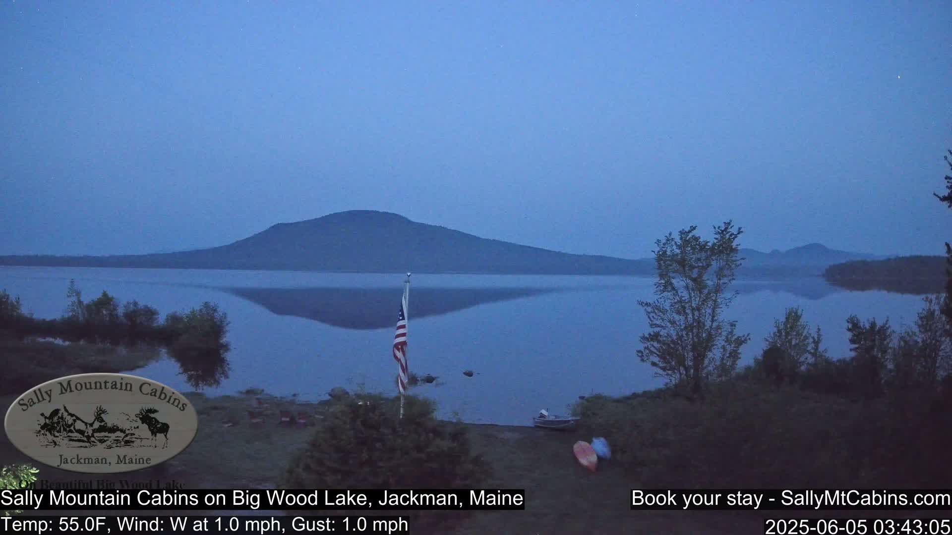 A calm, twilight scene shows a lake reflecting a distant mountain under a clear, dusky blue sky.
