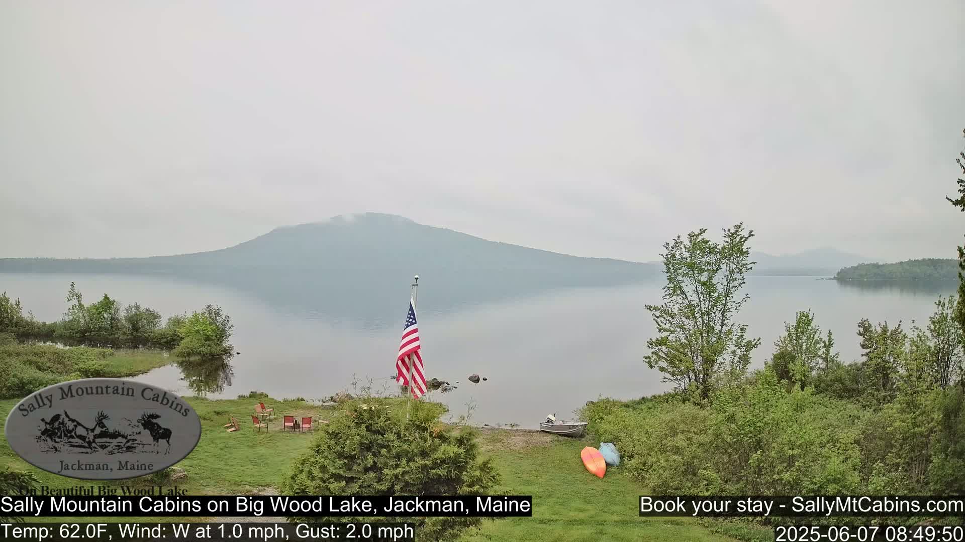 A hazy view of a calm lake, backed by a mountain range, with an American flag planted near a lakeside lawn containing some outdoor furniture and kayaks.