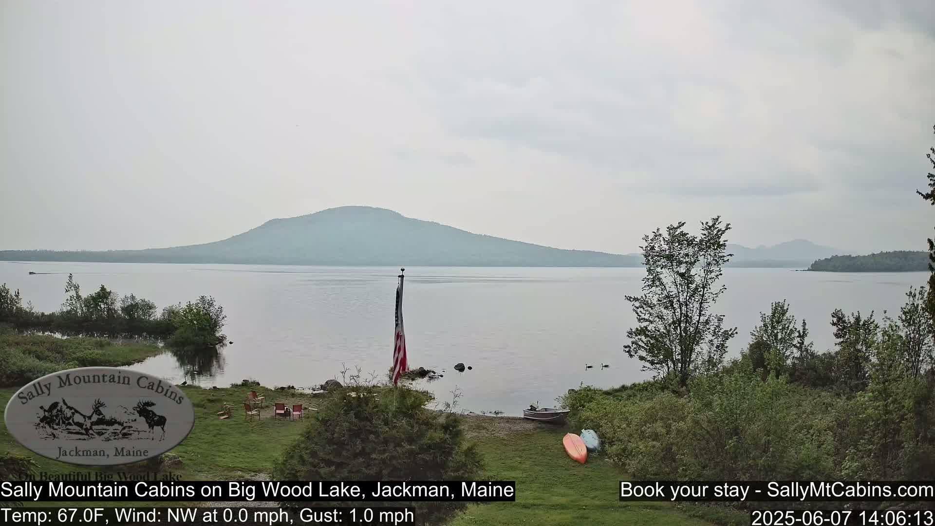 A calm lake with a mountain in the background is viewed from a grassy shore with a flagpole and a small boat; the sky is overcast.