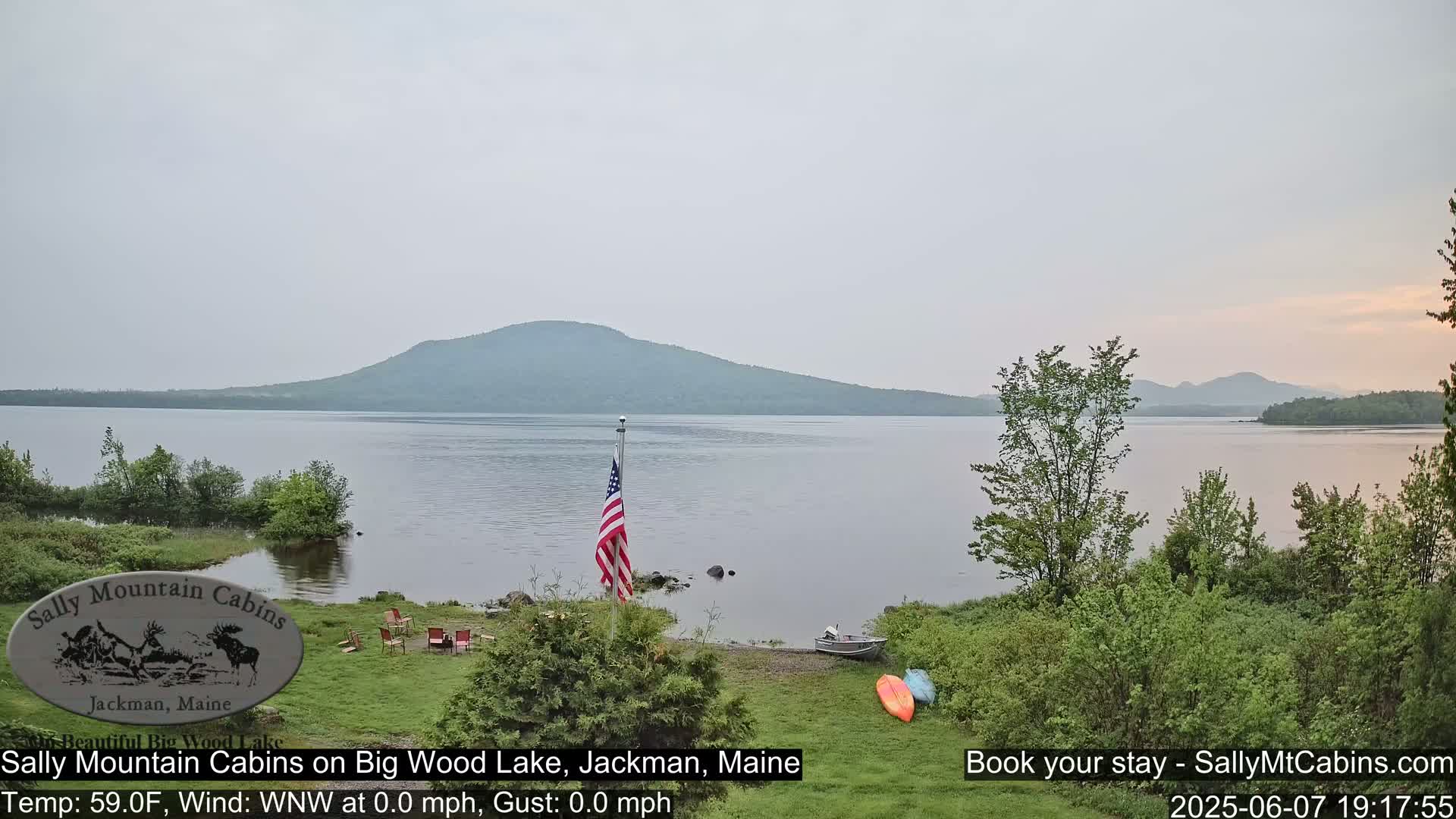 A calm lake with a mountain in the background is viewed from a lakeside lawn with an American flag and several recreational items.