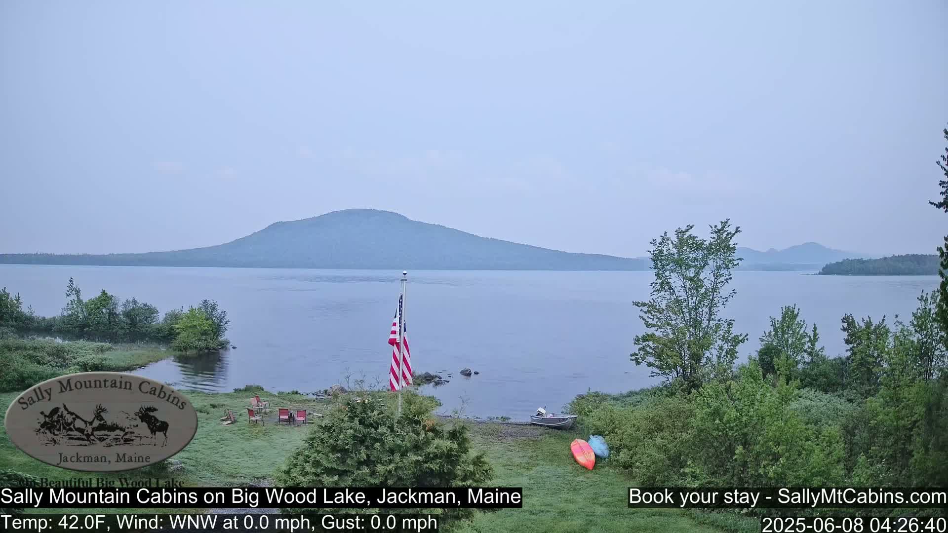 A calm, hazy day reveals a lake view with a distant mountain range, an American flag, and a small boat near a shoreline with green vegetation.