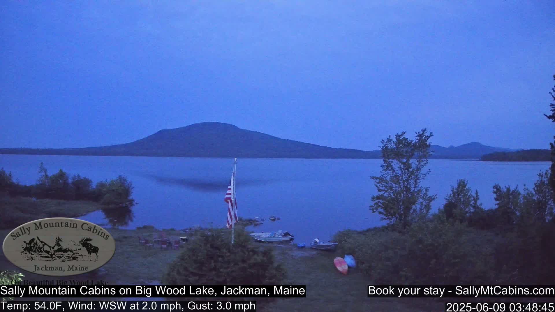 A calm, twilight view of a lake with a mountain in the background, boats on the shore, and an American flag.