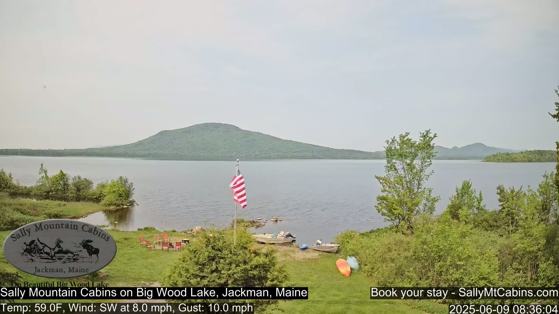 A calm lake with a wooded shore and distant mountains under a mostly cloudy sky, featuring several boats and kayaks pulled ashore near a flagpole and some outdoor seating.