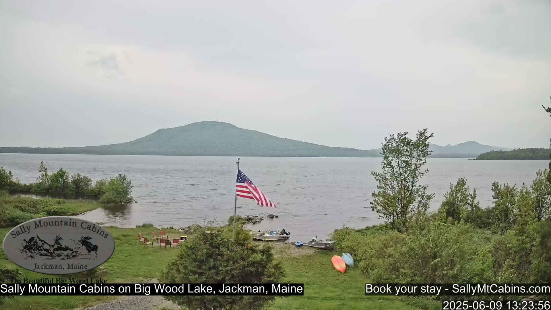 A calm lake with a gently sloping wooded hill in the background under an overcast sky, showing several boats and outdoor furniture on the lakeshore.