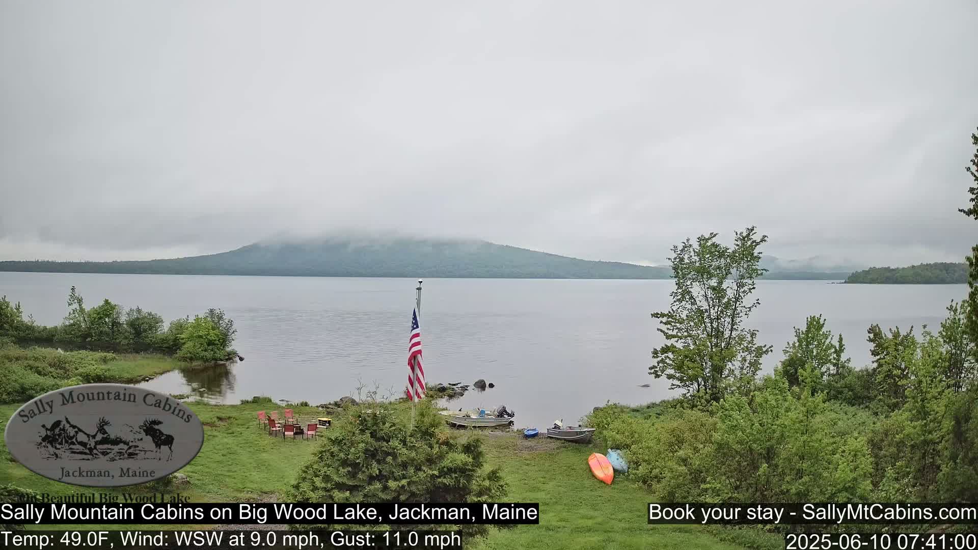 A calm lake with a misty mountain in the background is viewed from a lakeside campsite with boats and kayaks on the shore under an overcast sky.