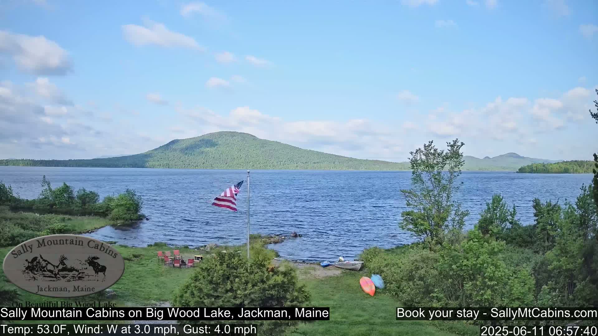 A calm lake with a forested mountain in the background, under a partly cloudy sky, is viewed from a lakeside location with some outdoor furniture and kayaks visible.